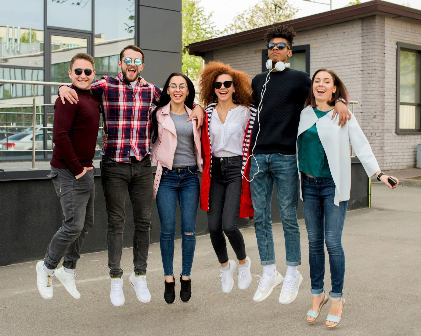 Group of six diverse, smiling college-age friends in stylish, casual outfits (jeans, sneakers, sunglasses) jumping in the air together outside a modern building, suitable for a blog about group picture day outfits or student fashion.