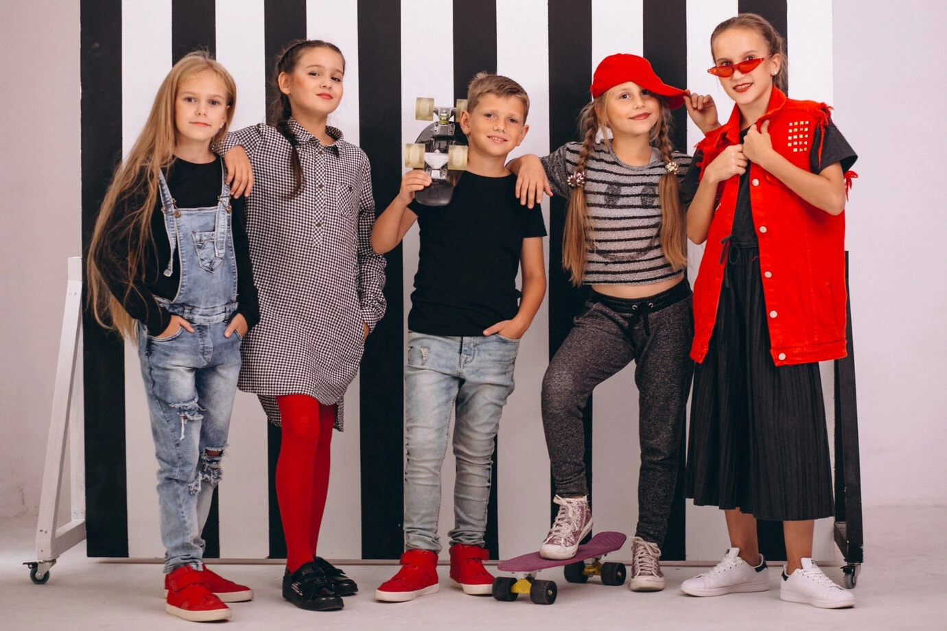 Group of five stylish kids and pre-teens posing in trendy picture day outfits against a black and white striped background, featuring distressed jeans, a red vest, and skateboards.
