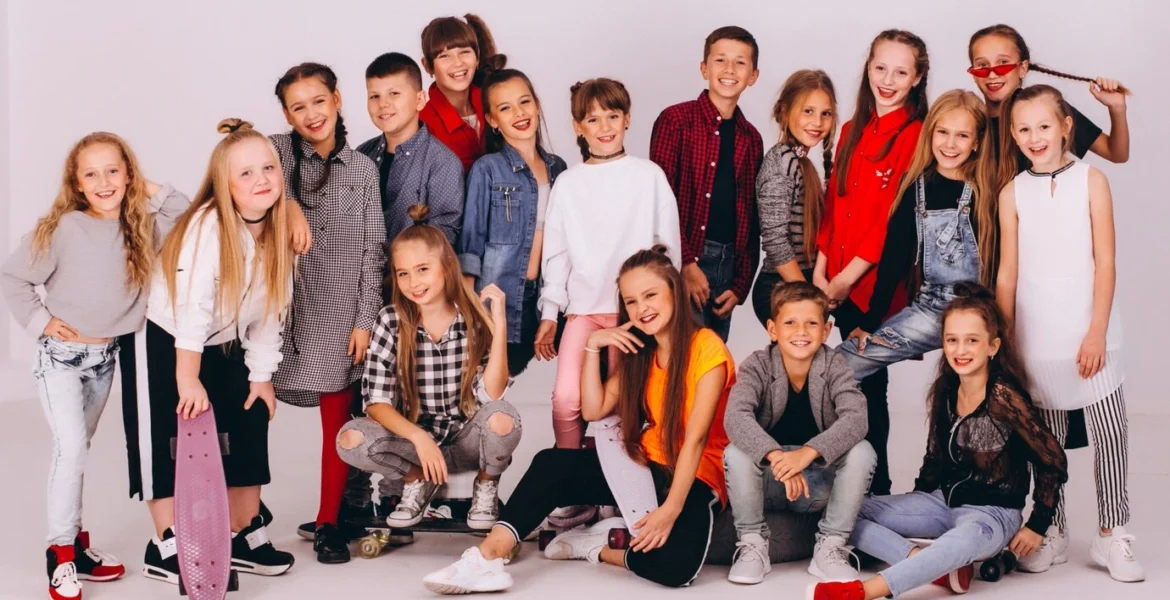 Large group of diverse and happy children and pre-teens posing for a photo in stylish, modern, and varied casual picture day outfits, including jeans, sweaters, and dresses, with some holding skateboards.