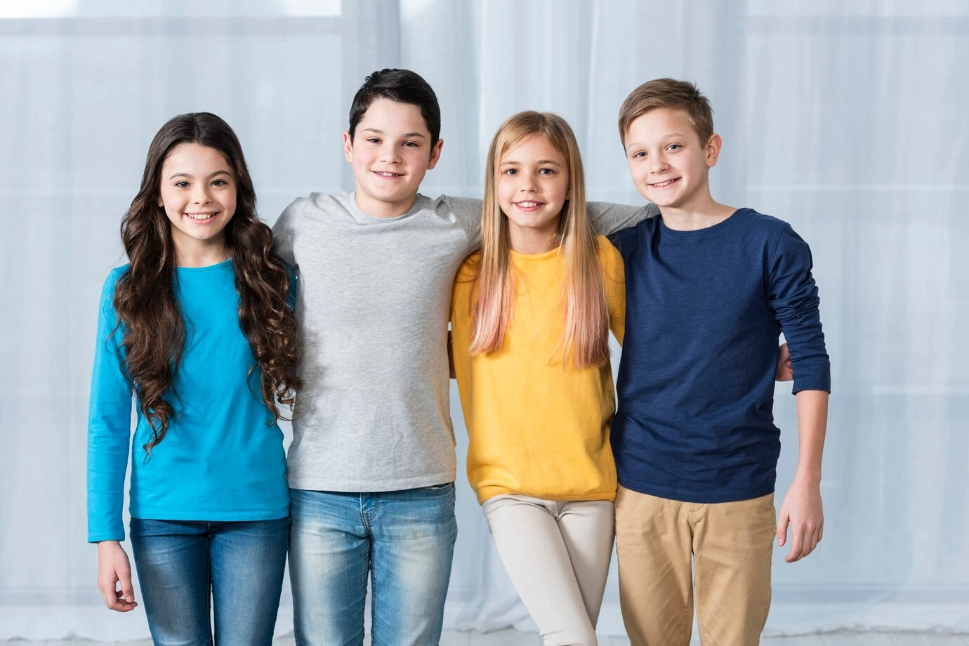 Four smiling kids and pre-teens with arms around each other, wearing simple, bright-colored picture day outfits like solid long-sleeve shirts and blue or tan jeans.