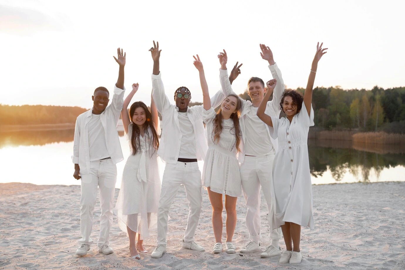 Group of six happy, diverse young adults dressed in matching all-white outfits (white dresses, shirts, and pants), standing on a beach with their hands raised in celebration at sunset, suitable for themes like unity or white party fashion.