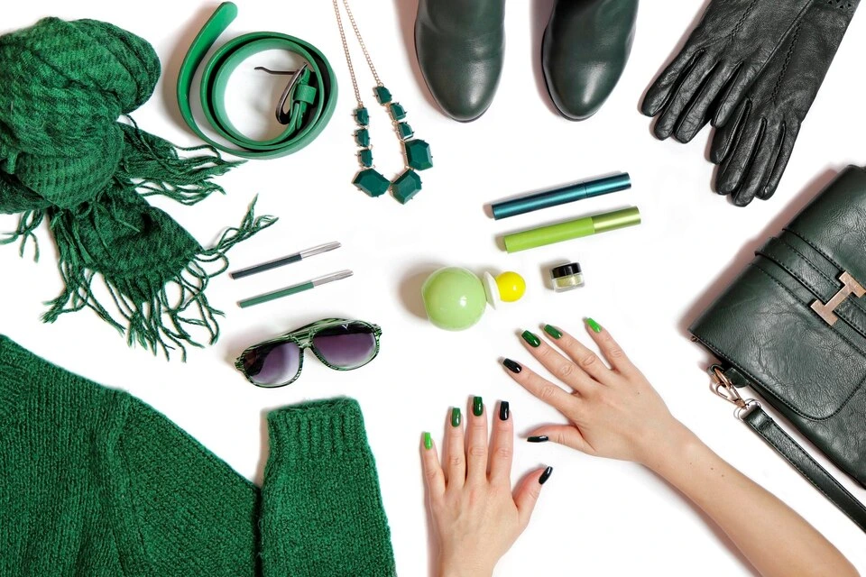 Overhead flat lay of various green fashion accessories and beauty products, including a chunky knit scarf, leather belt, necklace, ankle boots, black leather gloves, a handbag, sunglasses, and hands with green and black nail polish.